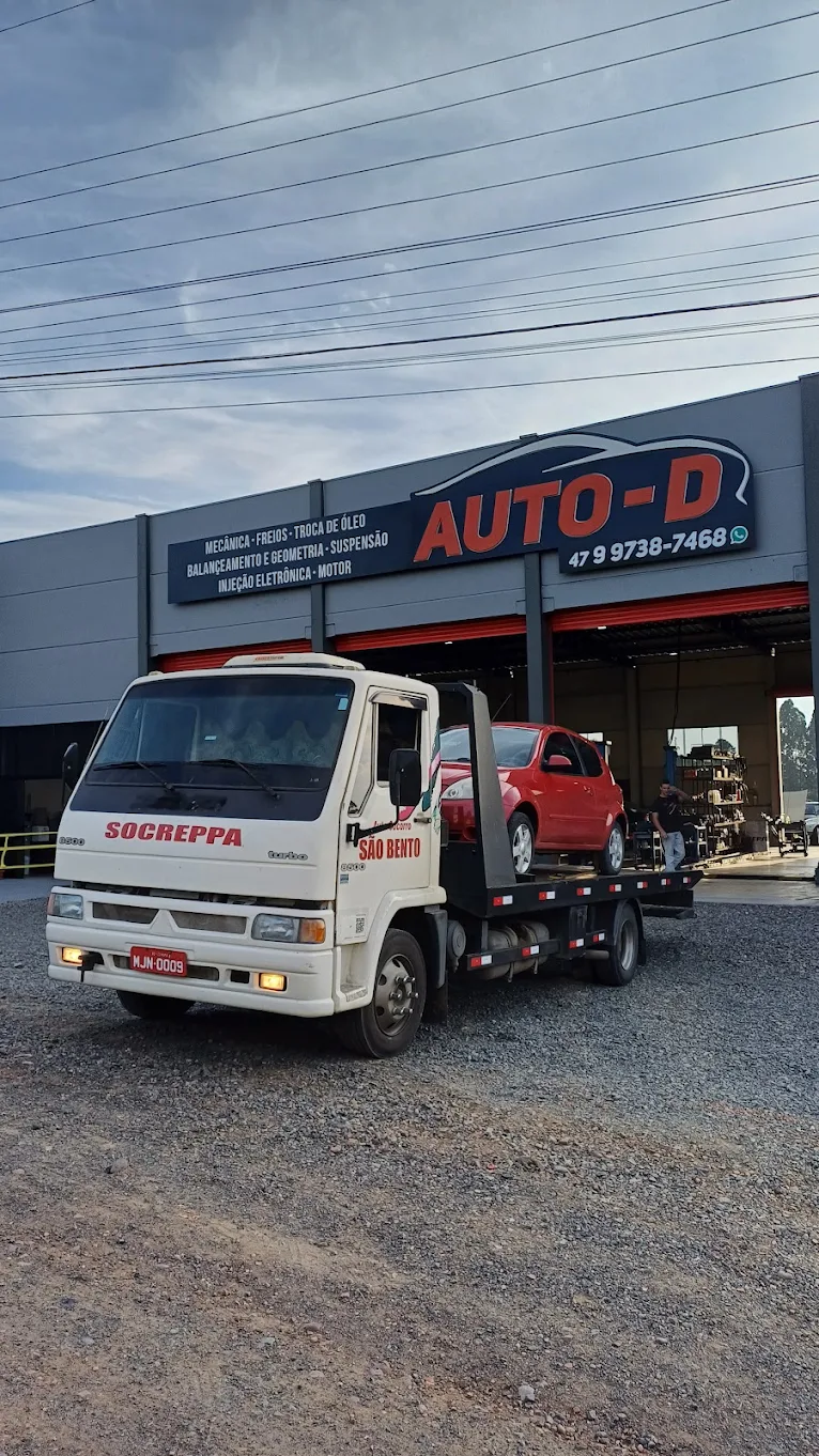 Atendimento da Auto-D Centro Automotivo com veículo sendo entregue por guincho na entrada da oficina, com painel de serviços ao fundo